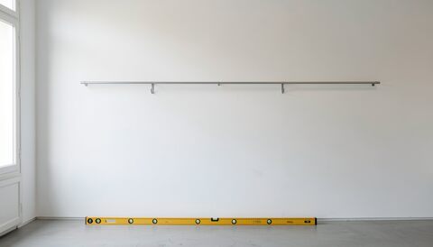 Mur blanc nu d'une salle d'exposition portant un rail d'accrochage en métal fixé par des équerres, niveau à bulle jaune posé au sol contre la plinthe, sol gris clair, lumière rasante venant d'une porte vitrée à gauche, aucun tableau.